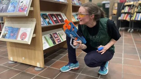 Image of a woman in hiding behind a book shelf at a library with nerf gun.